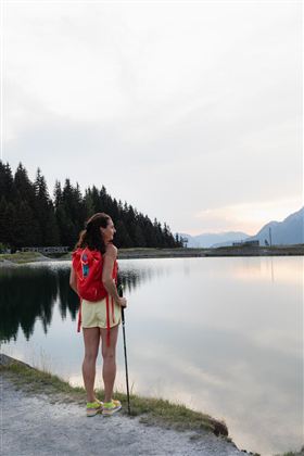 Een vrouw staat aan de oever van een kalme meer en kijkt in de verte. Op de achtergrond zijn groene bossen en bergen te zien.