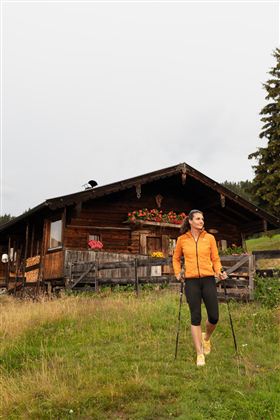 Een vrouw in een oranje jas wandelt op een weiland. Op de achtergrond staat een houten huis met bloembakken.