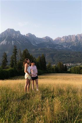 Twee personen staan op een groene weide met uitzicht op majestueuze bergen op de achtergrond. Het is een zonnige dag en het landschap straalt een rustige sfeer uit.