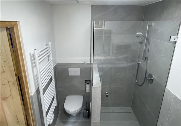 Een moderne badkamer met een glasdouche en grijze tegels. Rechts bevindt zich een toilet en een handdoekradiator.