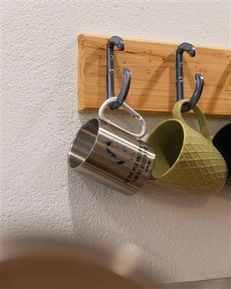 Een houten plank met twee haken, waaraan een zilveren en een groene mok hangen. De mokken hebben verschillende ontwerpen en zijn aan een witte muur geplaatst.