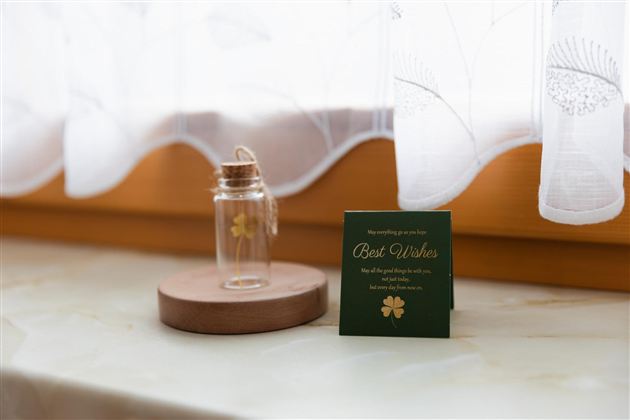 Een glas met een gouden klavertje vier staat op een houten onderzetter. Ernaast ligt een kaart met de tekst "Best Wishes".