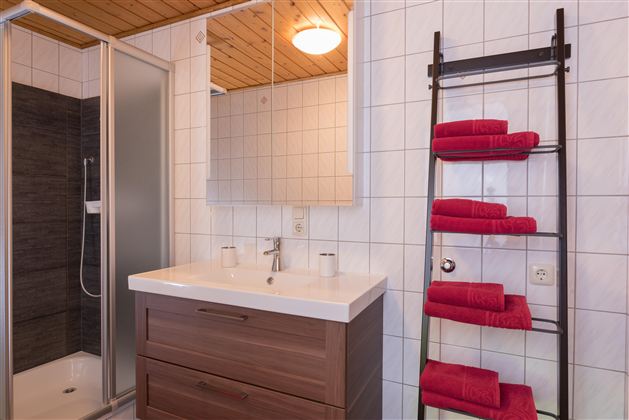 Ein modernes Badezimmer mit einer Dusche, einem Waschbecken und einem großen Spiegel. Rote Handtücher sind ordentlich an einem schwarzen Regal gestapelt.