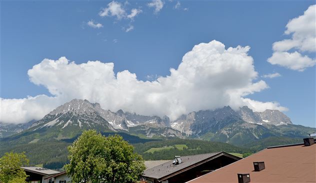 Eine beeindruckende Berglandschaft mit großen, bewaldeten Hängen und schneebedeckten Gipfeln. Der Himmel ist blau, und weiße Wolken ziehen vorbei.