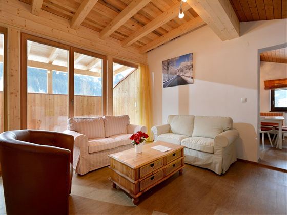 Ein gemütliches Wohnzimmer mit Holzdecke und großen Fenstern. Es gibt bequeme Sofas und einen Holztisch mit einem Blumenarrangement.