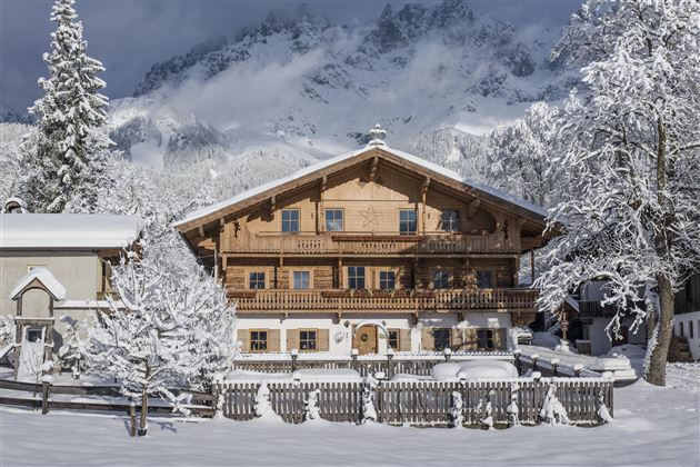 A charming wooden house in the snow, surrounded by snow-covered trees. Majestic mountains are visible in the background.