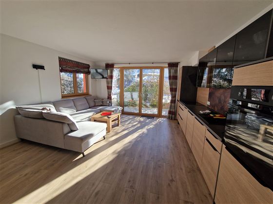 Ein modernes Wohnzimmer mit einem großen Sofa und einem Holztisch. Große Fenster lassen viel Tageslicht herein und bieten einen schönen Blick nach draußen.