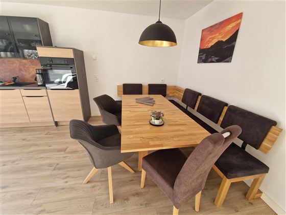 A modern dining area with a large wooden table and comfortable chairs. In the background, an elegant kitchen and a piece of artwork on the wall can be seen.