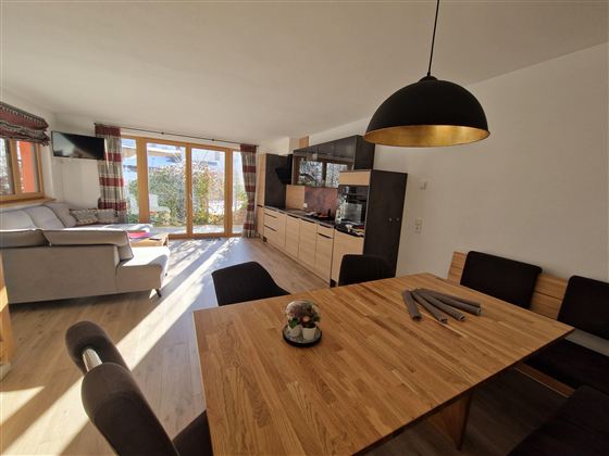 A modern living room with an open kitchen. A large wooden dining table and a cozy seating area by the window are clearly visible.