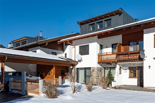 Een modern huis in de sneeuw met een houten balkon en een helderblauwe lucht. De omgeving lijkt rustig en uitnodigend.