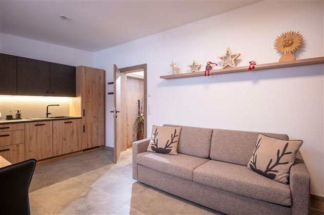 Een moderne woonkamer met een beige bank en een houten wandplank. De keuken op de achtergrond is van hout en goed verlicht.