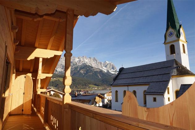 Ein Balkon mit Holzverkleidung und Blick auf eine malerische Kirch und majestätische Berge. Der Himmel ist klar und die Architektur ist traditionell.