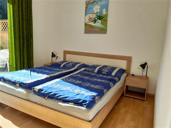 Ein gemütliches Schlafzimmer mit einem Doppelbett und blauen Bettdecken. An der Wand hängt ein Gemälde und es gibt einen Nachttisch mit einer Lampe.