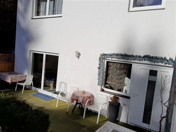 Eine Terrasse mit weißen Stühlen und Tischen. Der Hintergrund zeigt eine weiße Wand und Fenster.