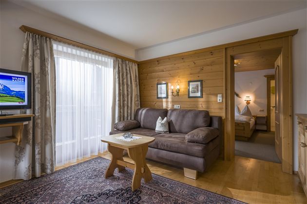 A cozy living ambiance with a brown sofa and a small table. The room has large windows with curtains and access to another room.