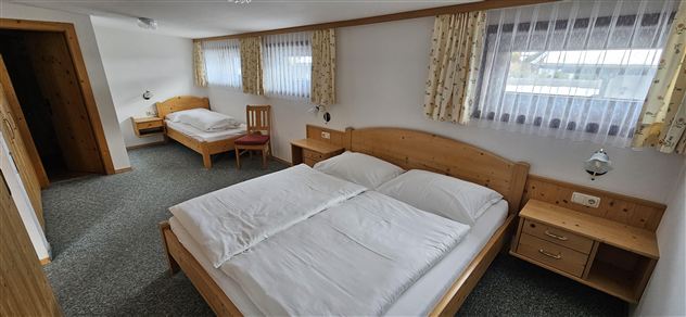 A simple guest room with two beds and a large window. The walls are bright and there is wooden furniture.