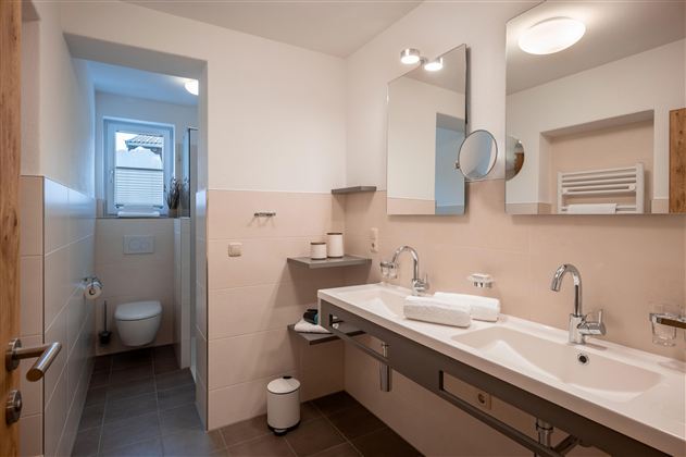 Moderní koupelna s dvojitým umyvadlem, velkým zrcadlem, sprchou, WC a ohřívačem ručníků.
