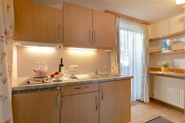 A modern kitchen with wooden cabinets and a sink. A window curtain lets light in, and on the countertop, there is a teapot and fresh fruit.