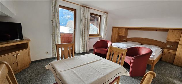 A cozy room with a double bed, two red chairs, and a table. Large windows offer a view of the mountains outside.