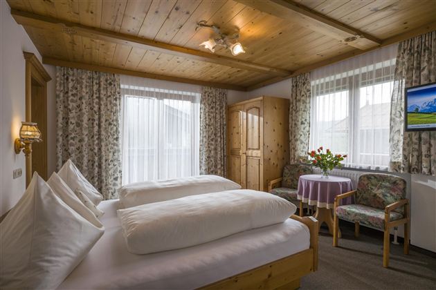 A cozy bedroom with two beds and a wooden ceiling. Daylight streams through the windows, and a table with flowers creates a pleasant atmosphere.