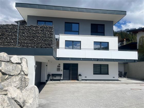 Ein modernes Gebäude mit mehreren Fenstern und einem Balkon. Die Fassade ist in Weiß und Grau gehalten, umgeben von einem Parkplatz und einer steinernen Mauer.