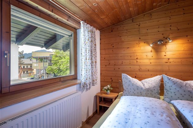 A cozy bedroom with wooden paneling and a window overlooking a green view. The beds are invitingly decorated with white pillows and blankets.