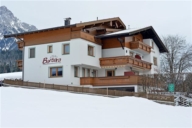 Ein gemütliches Haus im alpinen Stil, umgeben von Schnee. Im Hintergrund sind Berge sichtbar, was eine malerische Winterlandschaft schafft.