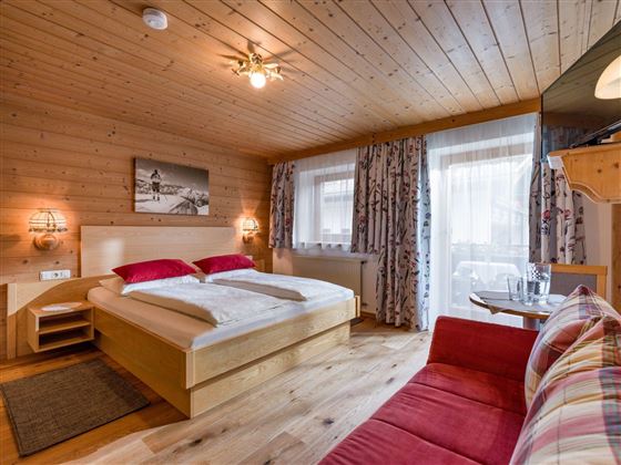 Ein gemütliches Schlafzimmer mit Holzverkleidung und großen Fenstern. Das Zimmer verfügt über ein Doppelbett, einen roten Sofa-Bereich und dekorative Vorhänge.