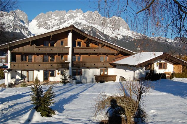 Ein skandinavisches Holzhaus im Schnee mit Bergen im Hintergrund. Die Szenerie ist ruhig und sonnig.