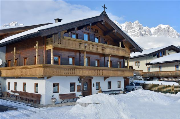 Ein traditionelles Chalet im Schnee mit einem großen Balkon und einem Kreuz auf dem Dach. Im Hintergrund sind schneebedeckte Berge zu sehen.