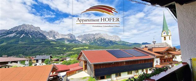 Eine malerische Aussicht auf Berge und ein dörfliches Panorama. Im Vordergrund sind Häuser und eine Kirche mit einem spitzen Turm zu sehen.