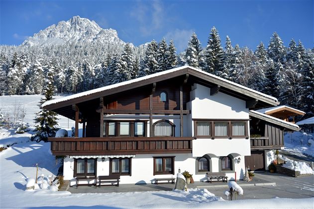 A beautiful chalet in the snow with a picturesque mountain backdrop. The surroundings are filled with many fir trees.
