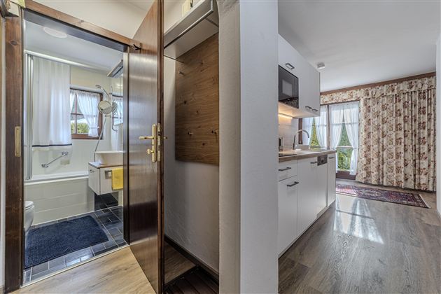 Ein modernes Badezimmer mit Dusche und hellen Vorhängen ist sichtbar. Daneben eine moderne Küche mit Holzregalen und Fenster mit Aussicht.