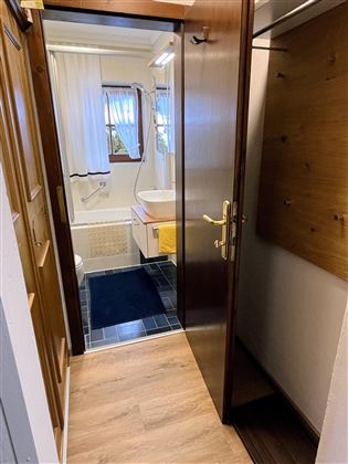 Ein Flur mit einem Blick auf ein Badezimmer. Die Wände sind hell und der Boden ist aus Holz, während eine Badewanne und eine Garderobe sichtbar sind.