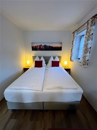 Ein gemütliches Zimmer mit Doppelbett und Nachttischlampen. Das Fenster hat eine florale Gardine und an der Wand hängt ein Bild.