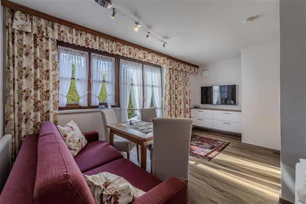 Ein gemütlicher Wohnbereich mit einem roten Sofa und einem Holztisch. Große Fenster lassen viel Licht herein und sorgen für eine einladende Atmosphäre.