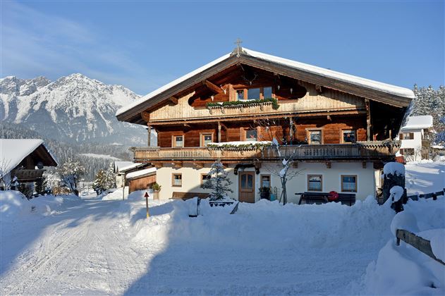 A traditional wooden house in the snow with mountains in the background. It is a clear, sunny day.
