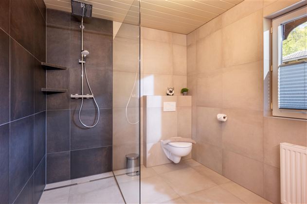 Ein modernes Badezimmer mit einer offenen Dusche und einer grauen Fliesenwand. Das WC ist einfach und funktional, mit einem Fenster, das Tageslicht hereinscheinen lässt.