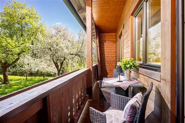 Ein gemütlicher Balkon mit Blick auf einen blühenden Garten. Ein Tisch mit Blumen und zwei Stühlen lädt zum Entspannen ein.