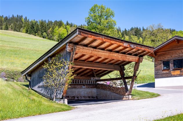 Eine moderne Garage aus Holz mit einem schrägen Dach steht in einer grünen Landschaft. Im Hintergrund sind sanfte Hügel und Bäume zu sehen.