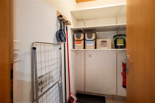 Ein kleiner Abstellraum mit Regalen und verschiedenen Aufbewahrungsboxen. An der Wand hängen Reinigungsutensilien und eine Garderobe.