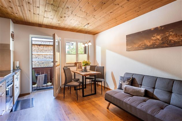 Eine helle und einladende Wohnküche mit einem Esstisch und einem gemütlichen Sofa. Große Fenster lassen viel Tageslicht herein und bieten einen Blick ins Freie.