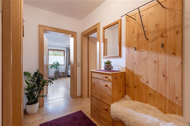 A bright entrance area with wooden cabinets and a mirror. In the background, plants and another room are visible.