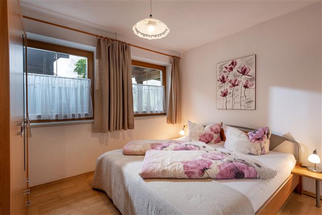 A cozy bedroom with a double bed and bright walls. Large windows and a floral painting create an inviting atmosphere.