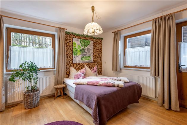 A cozy bedroom with a double bed, soft pillows, and natural wood elements. The walls are decorated with a stylish wallpaper, and large windows provide plenty of daylight.