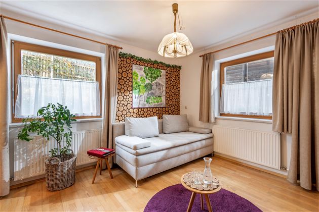 A cozy living room with a light couch and a round table on a purple rug. The room has large windows that let in plenty of daylight, and a decorative wall with plant motifs.