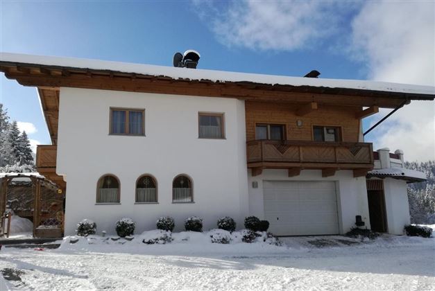 A cozy house in alpine style with a covered balcony. The surroundings are snow-covered and the sky is partly cloudy.