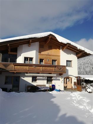 A beautiful wooden house in the snow with a balcony and a bright facade. The sky is blue and the area is surrounded by a tranquil winter landscape.