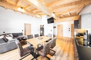 The Villaggio apartment offers a modern living-dining room with wooden ceilings, large windows, and a fireplace stove. There is a wooden dining table, cozy seating furniture, and access to a large balcony. On the balcony, there is a table with chairs inviting you to linger. Enjoy the view of the Wilder Kaiser and the village of Going am Wilden Kaiser.