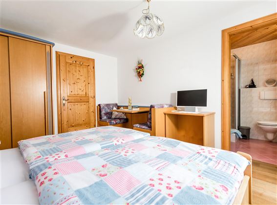 A cozy bedroom with a double bed and a wooden wardrobe. In the background, a small dining area and a bathroom are visible.